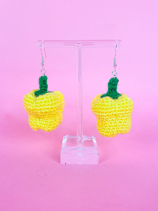 A pair of crochet earrings shaped like bell peppers in yellow attached to silver butterfly hooks hanging on an earring stand.