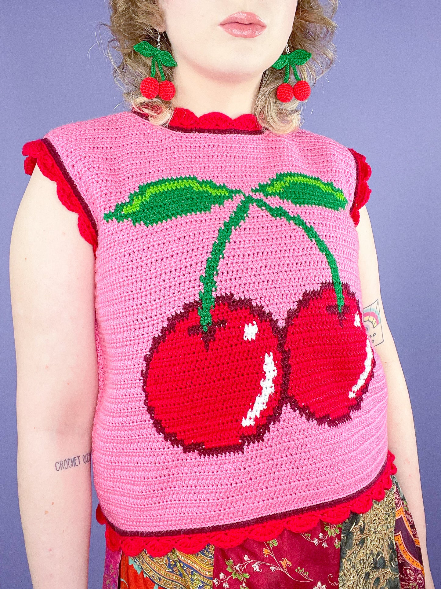 Close up of woman wearing a pink crochet vest with a red cherry on the front and frilly edges with matching cherry earrings.