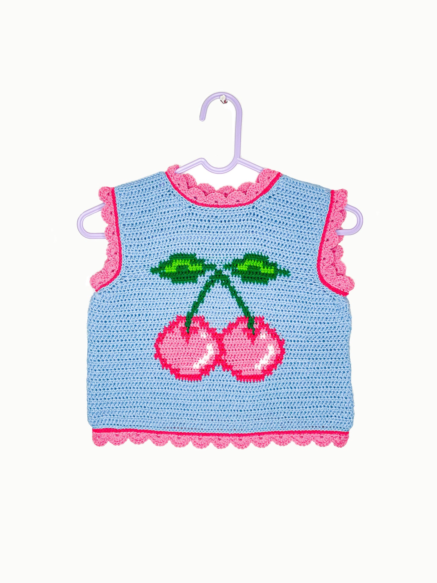 A baby blue crocheted vest with a pink cherry on the front and pink frilly edges, hanging on a white wall.