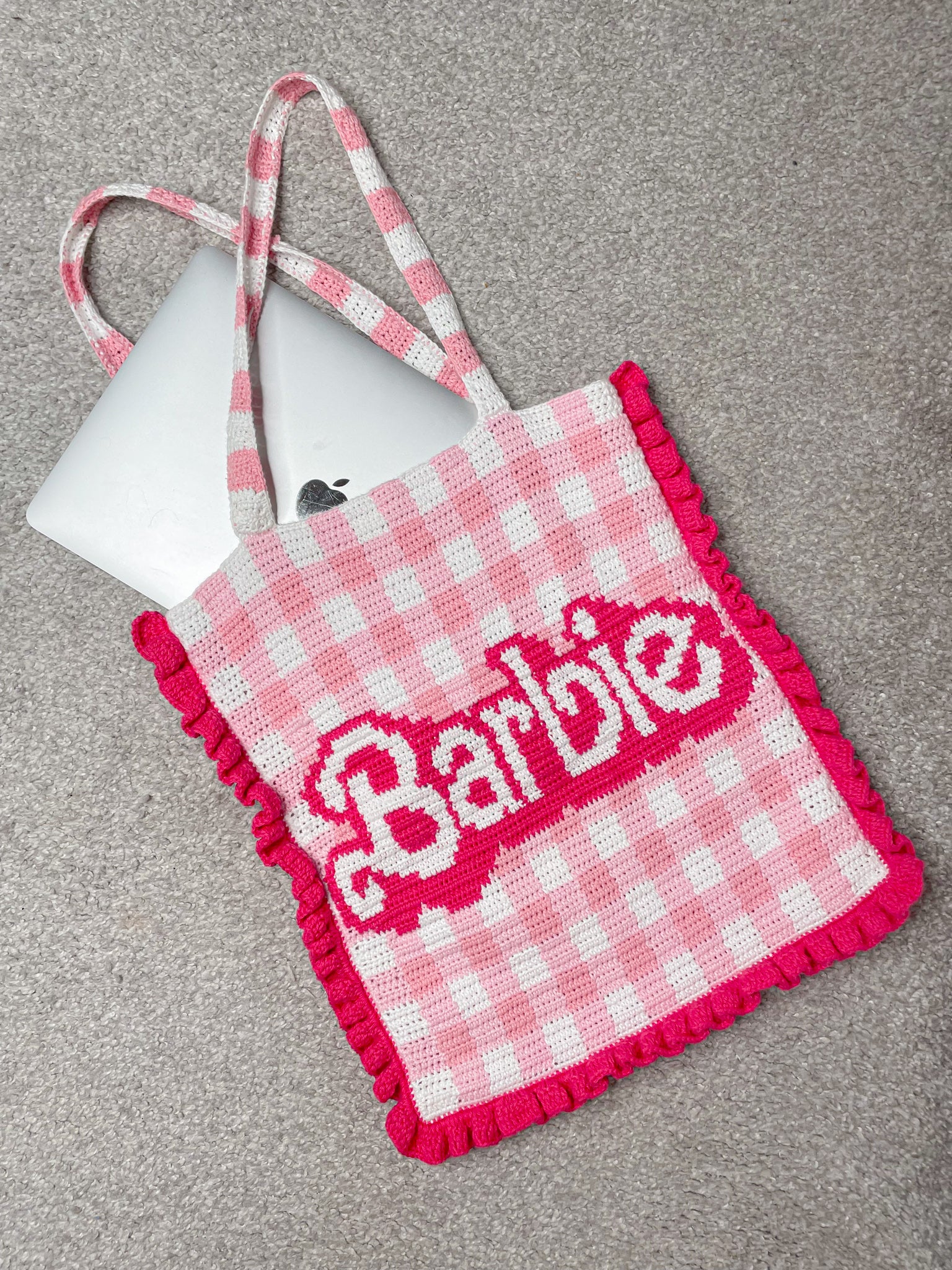 Flat lay of a barbie bag in pink and white with dark pink frills lying on a grey carpet with a MacBook hanging halfway outside the bag.