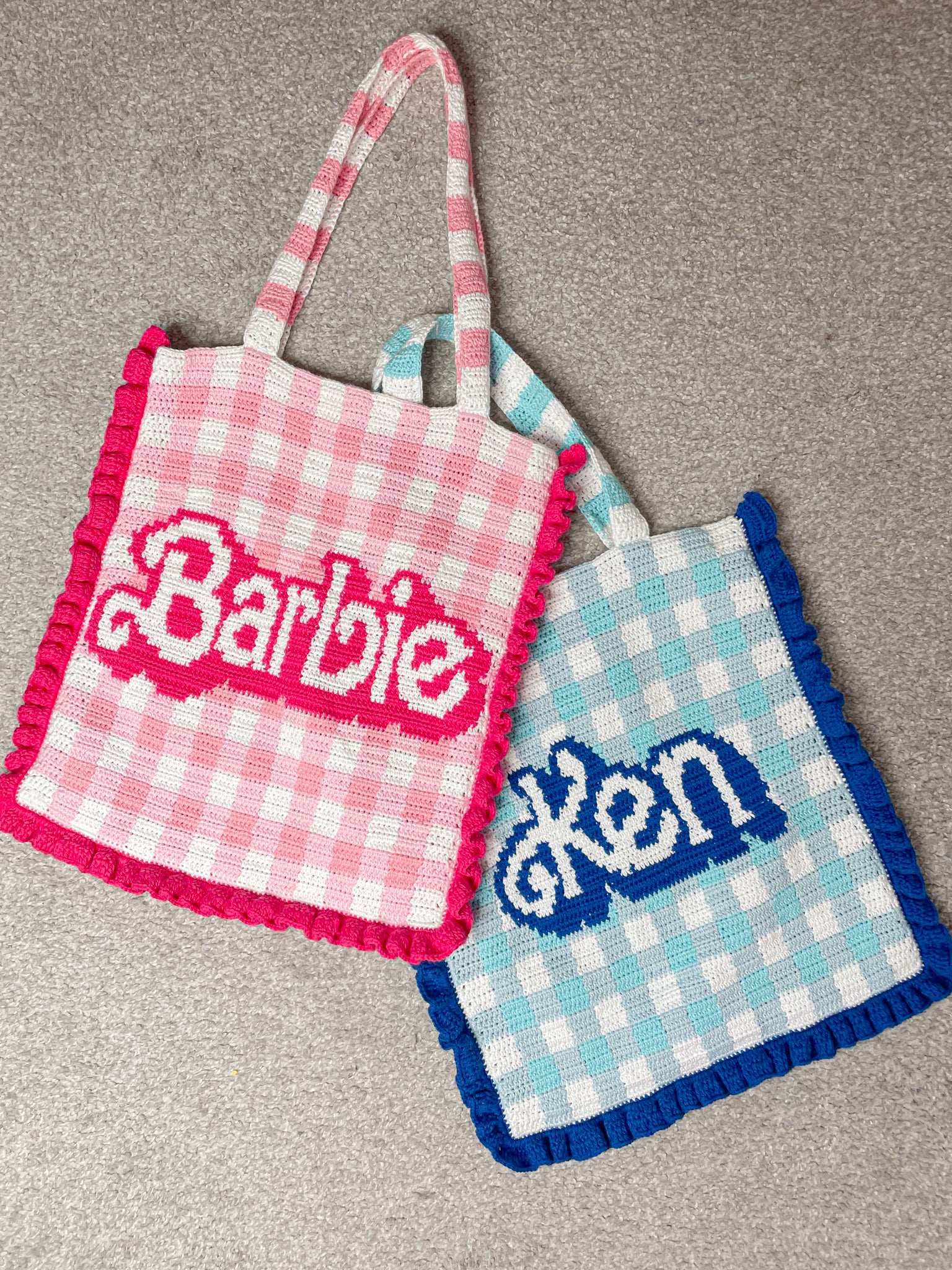 Flat lay of two tote bags, one in pink and white gingham with 'Barbie' on the front, the other in blue and white gingham with 'Ken' on it, lying together on a grey carpet.