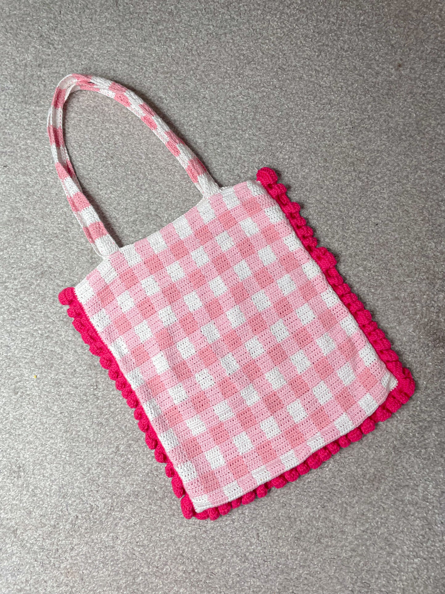 Flat lay back view of a pink and white gingham tote bag with dark pink frilly edges lying on a grey speckled carpet.