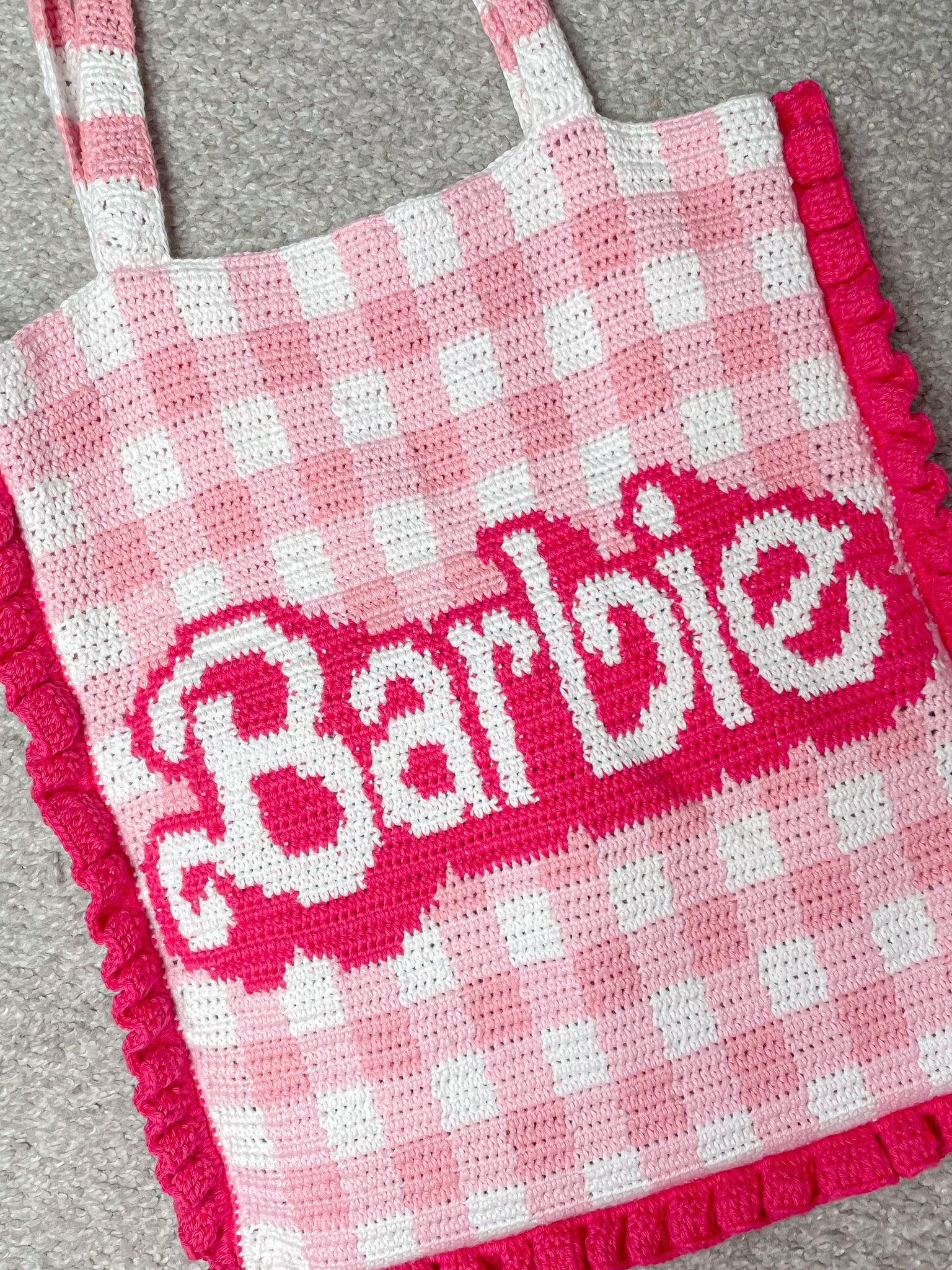 Flay lay close view of a crochet tote bag with a pink and white gingham design and the word 'Barbie' on the front lying on a light grey rug.