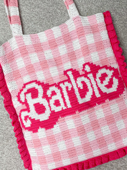 Flay lay close view of a crochet tote bag with a pink and white gingham design and the word 'Barbie' on the front lying on a light grey rug.