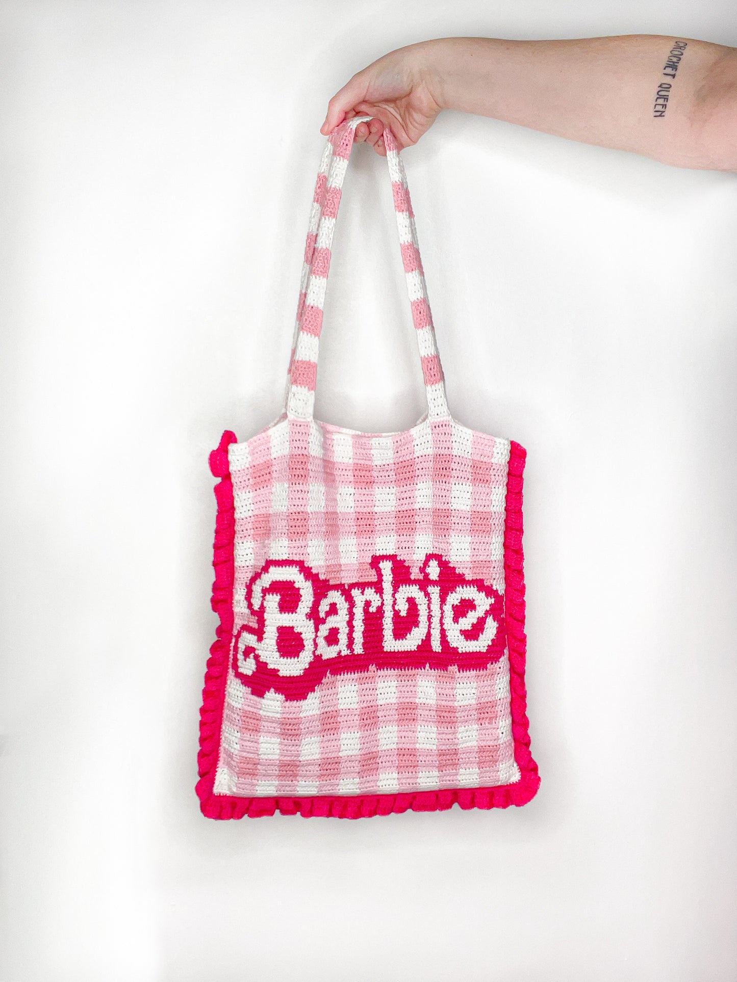 A hand holding a crochet bag in pink and white gingham with 'Barbie' on the front and a dark pink frilly edge held in front of a white wall.