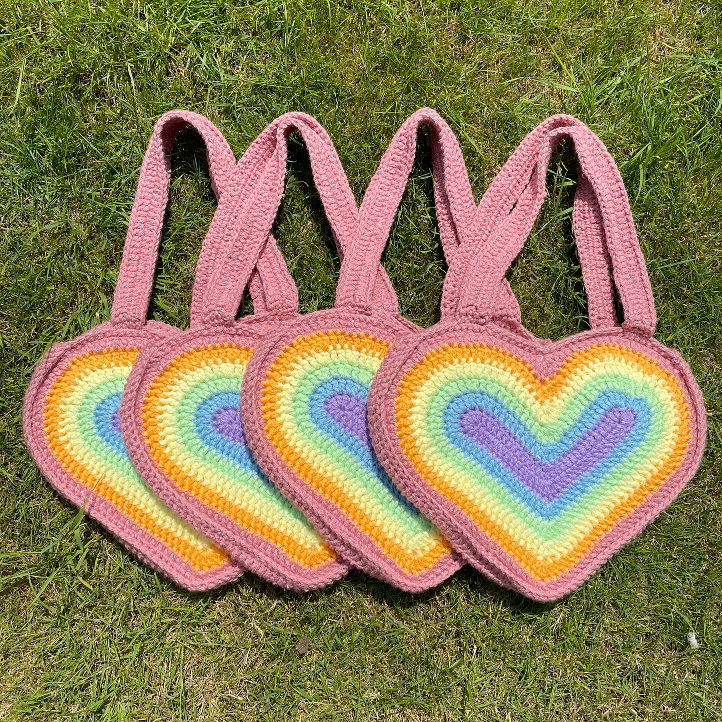 Four crochet bags shaped like hearts in pastel rainbow colours, lying together in a row on a patch of grass.