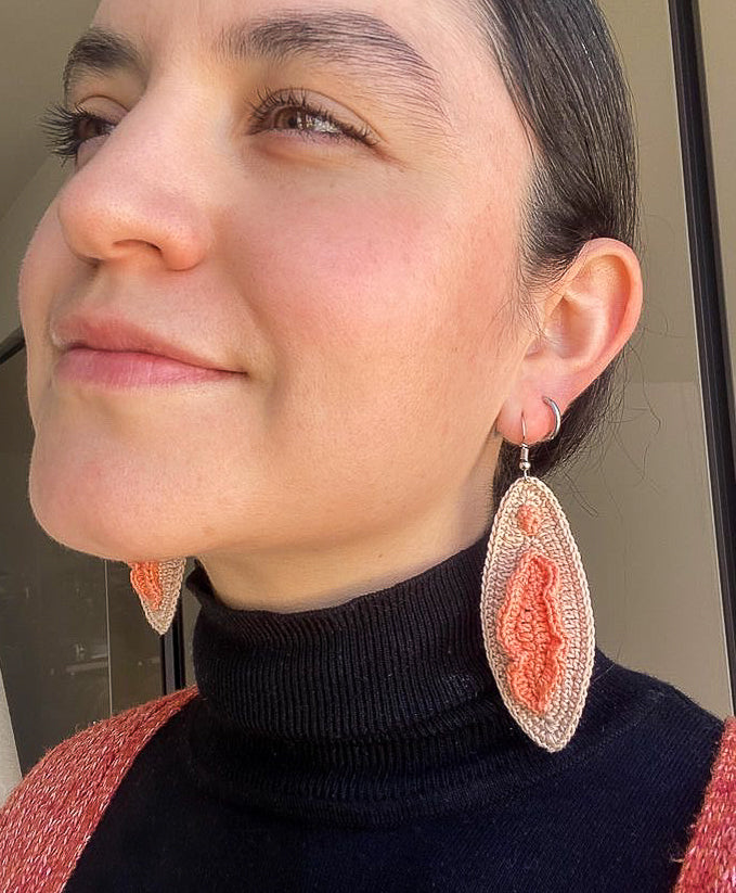 Close up of woman wearing a pair of crochet vulva drop earrings in brown and peach with a turtleneck and matching peach top.