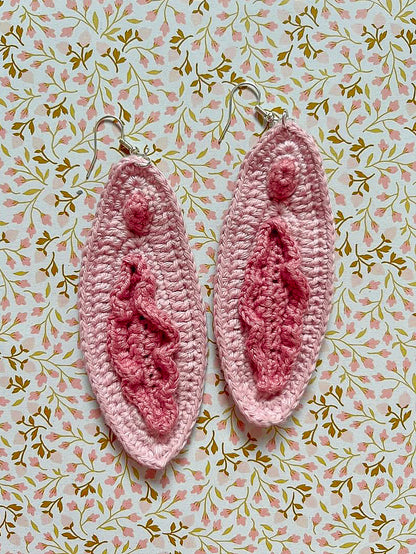 Flat lay of a pair of drop earrings in cream and pink that look like vulvas lying on a floral pink and brown background.