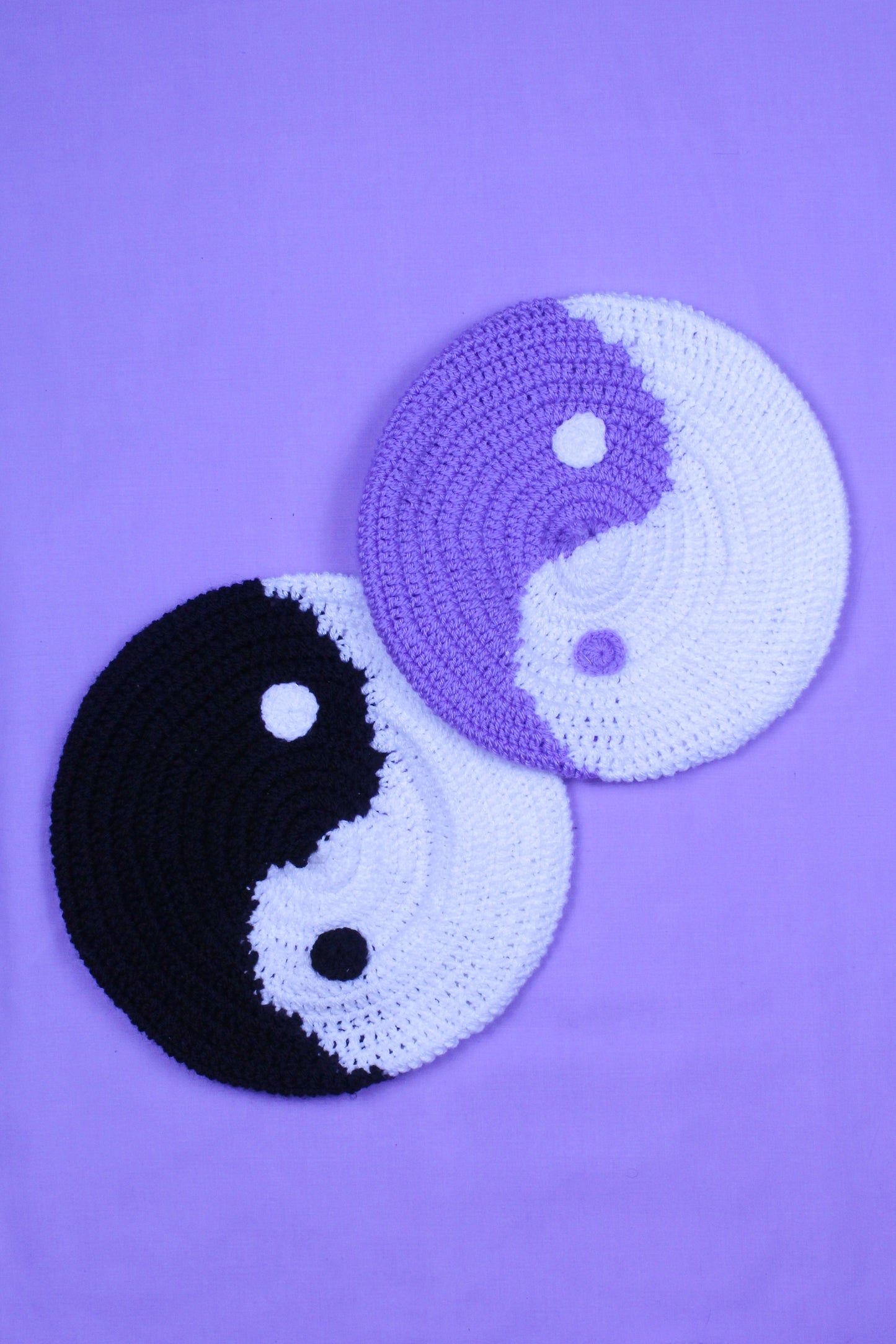 Two crocheted berets that look like the yin and yang symbol lying together, one in black and white and one in lilac and white.