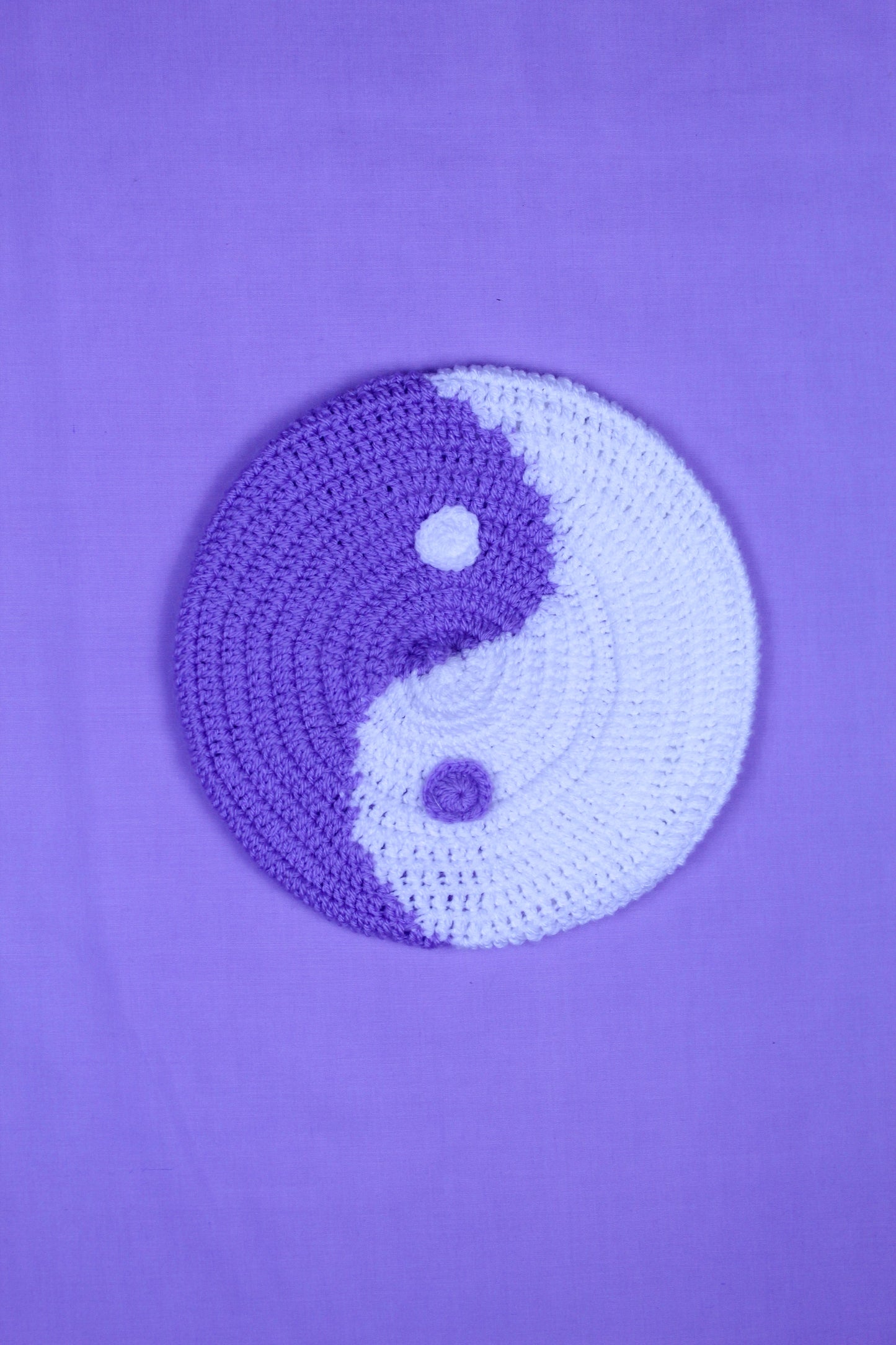 A purple and white beret with a yin and yang design lying on a purple background.