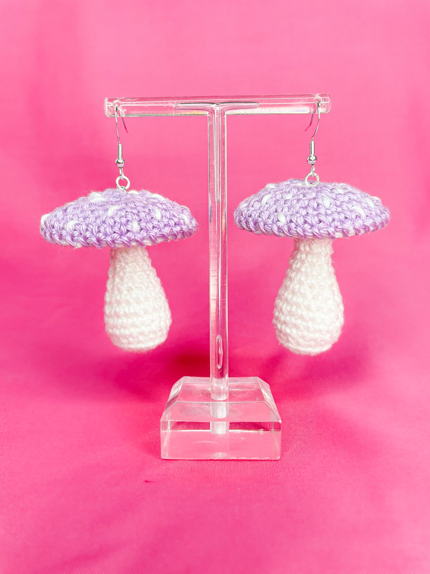 A pair of toadstool mushroom earrings in lilac attached to silver butterfly hooks hanging on an earring stand.