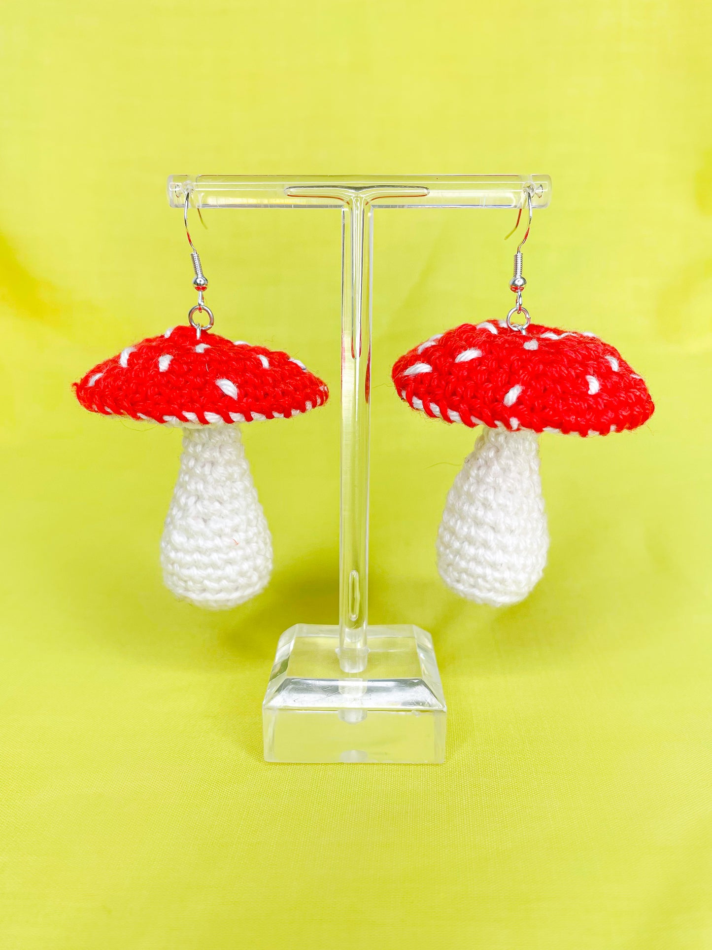 A pair of red and white crochet earrings shaped like mushrooms hanging on an earring stand with a yellow background behind them.