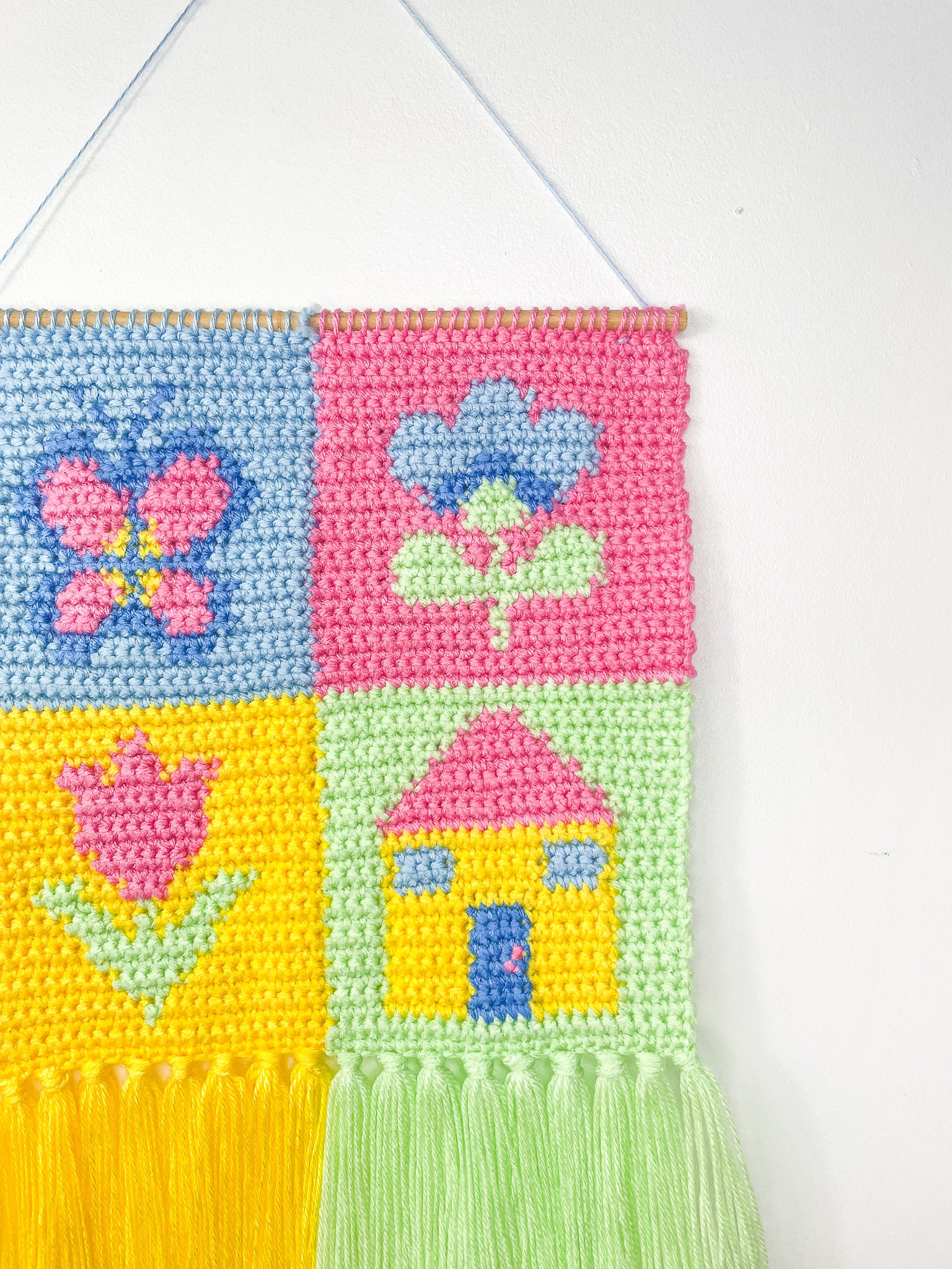 Side view close up of a patchwork crochet wall hanging in pastel colours hung up on a white wall and attached to a wooden dowel rod to keep its shape.