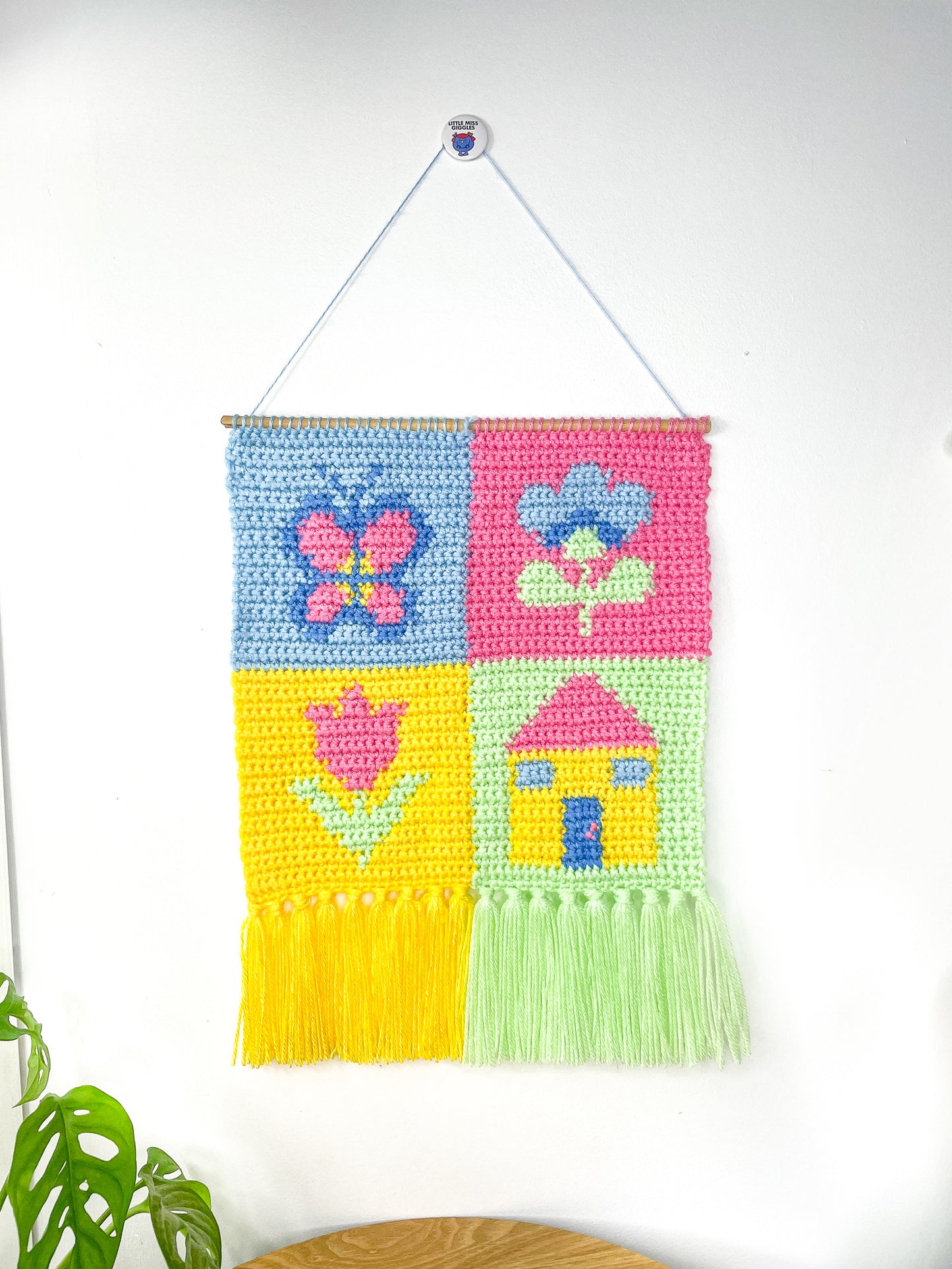 A patchwork pastel wall hanging with different images in each square (butterfly, two flowers and a house), hung on a white wall with a plant and wooden coffee table under it.