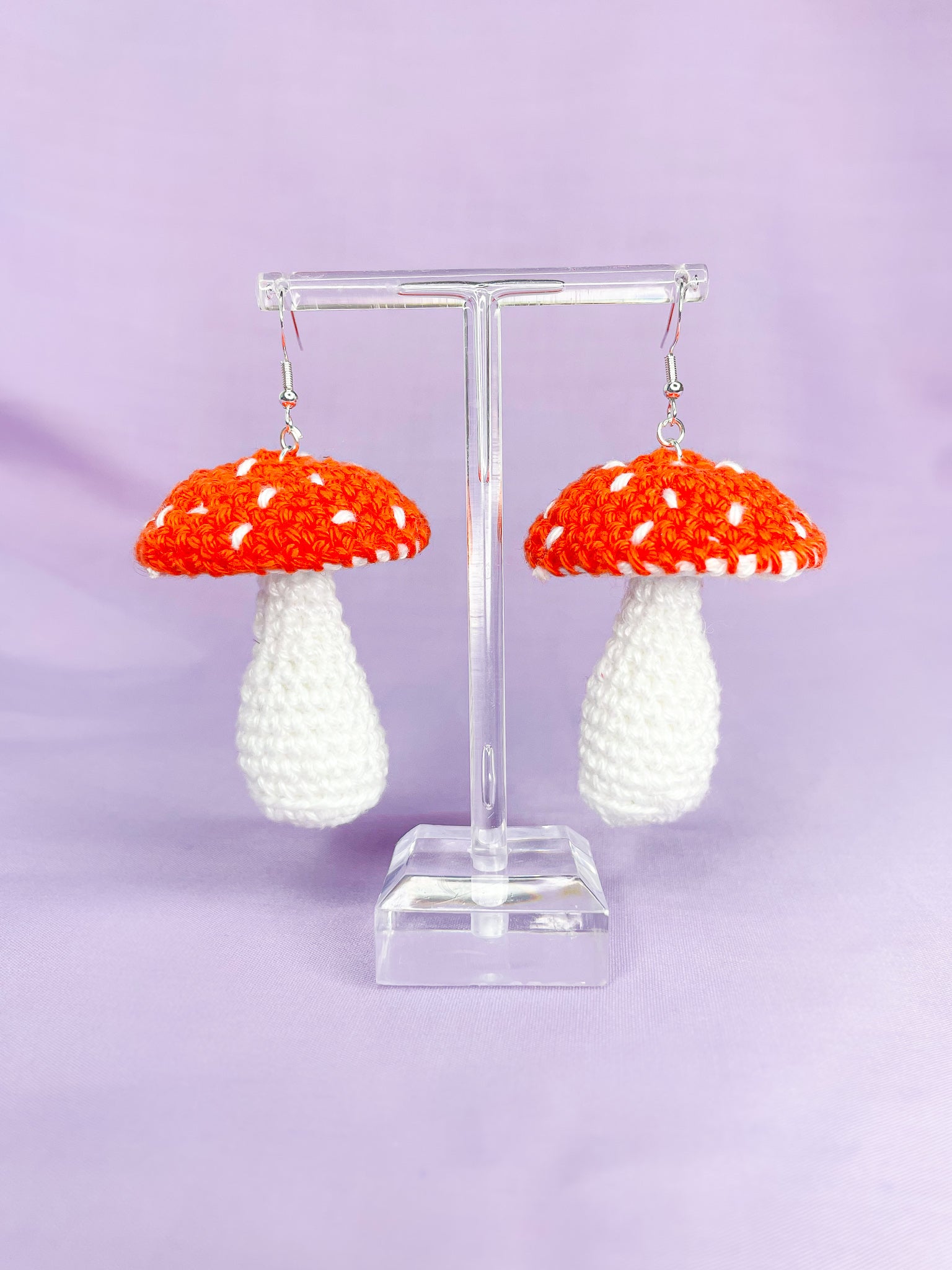 Pair of orange mushroom shaped earrings hanging on an earring stand with a lilac background behind it.