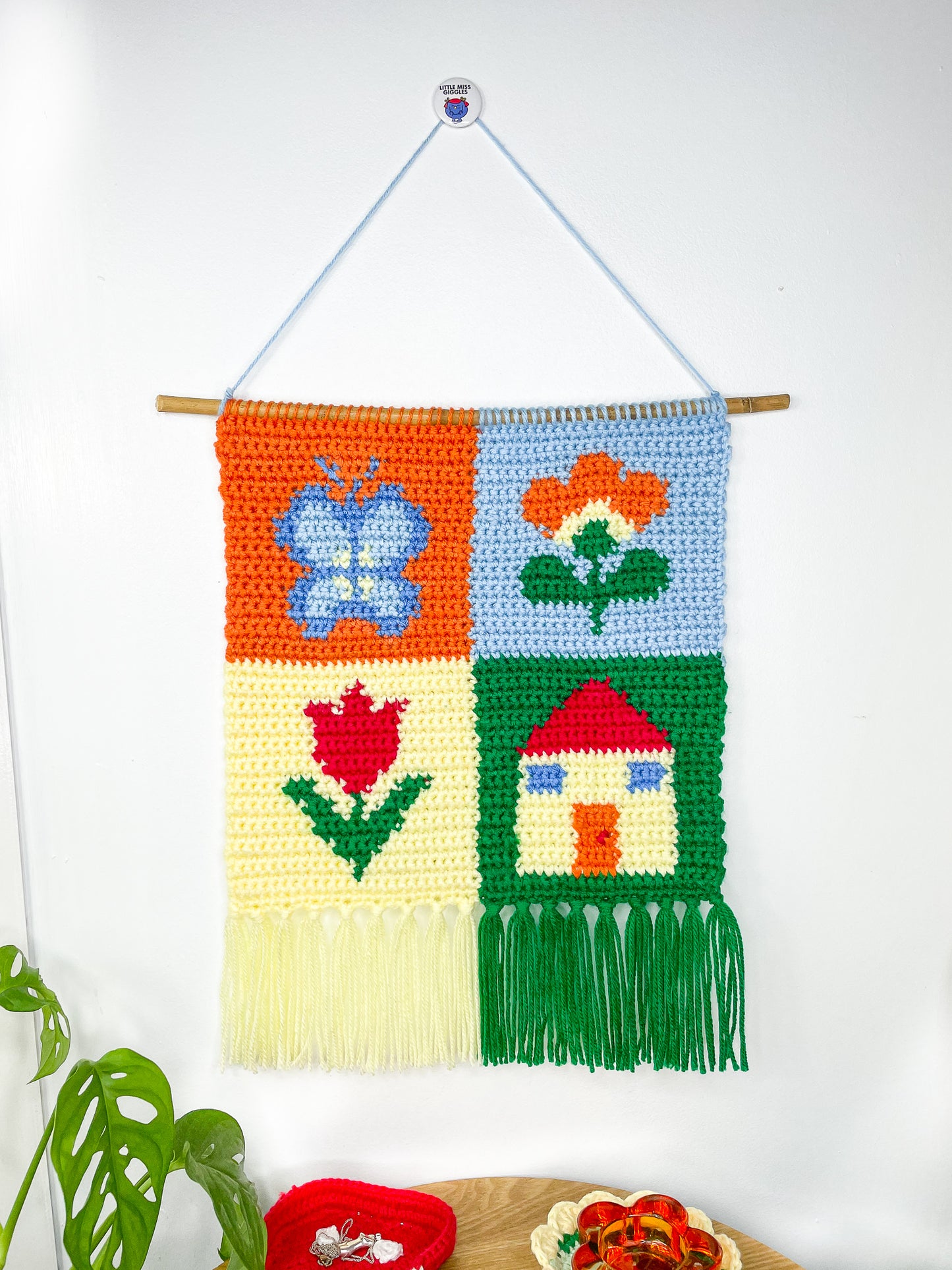 A crochet wall hanging with a patchwork design (orange, blue, pale yellow, green and red) and different images in each square (butterfly, house and two flowers) hung on a white wall.