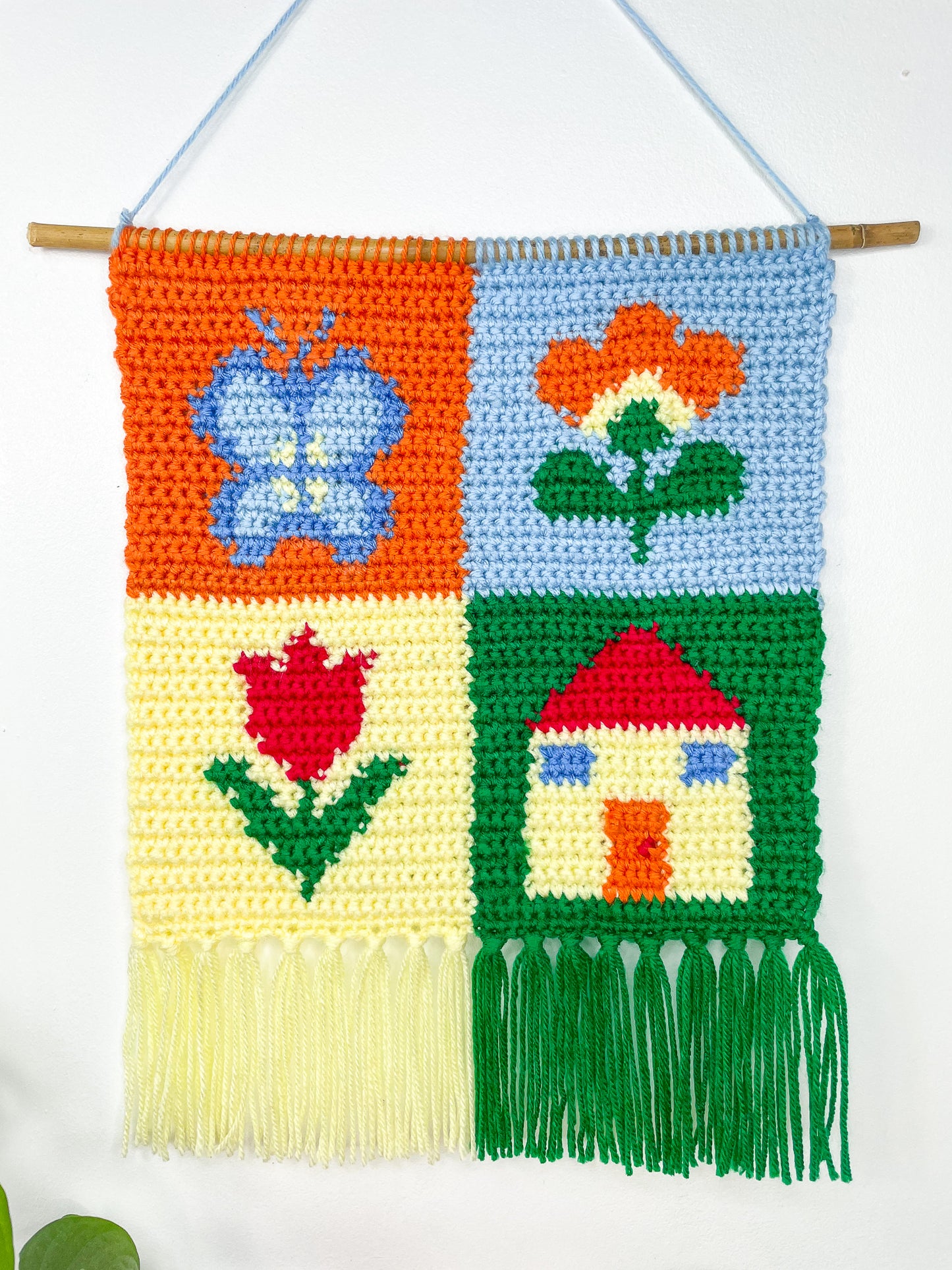 Close view of a crochet wall hanging on a white wall sewn to a wooden stick to keep its shape with a patchwork design and tassels attached to the bottom.