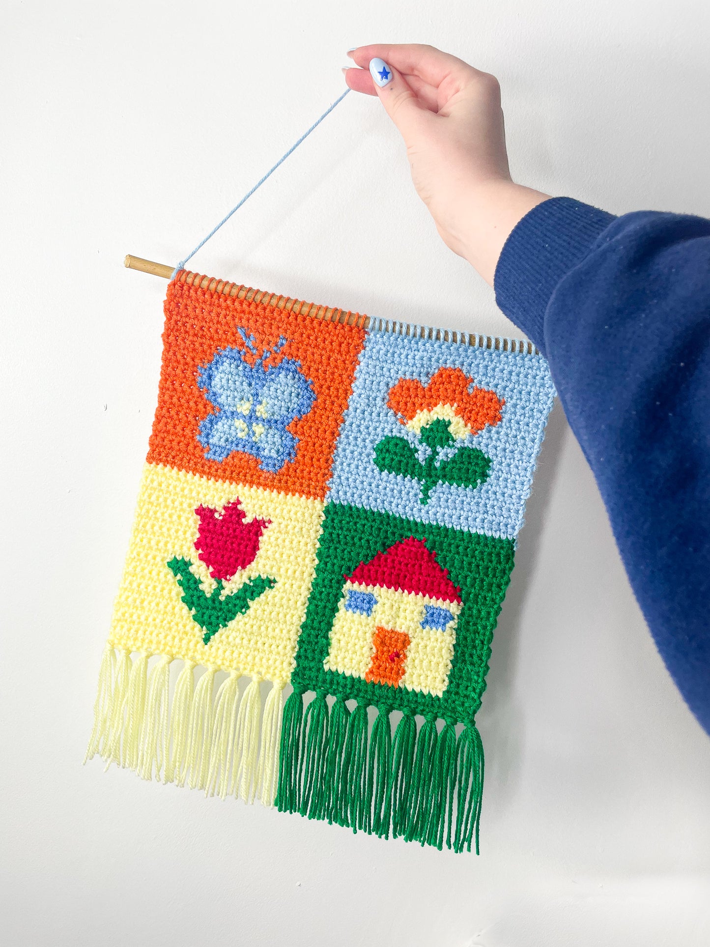 A hand holding a crochet tapestry with a patchwork design and four different coloured squares (orange, baby blue, pale yellow, emerald green), each square with a different image in it (butterfly, two flowers and a house).