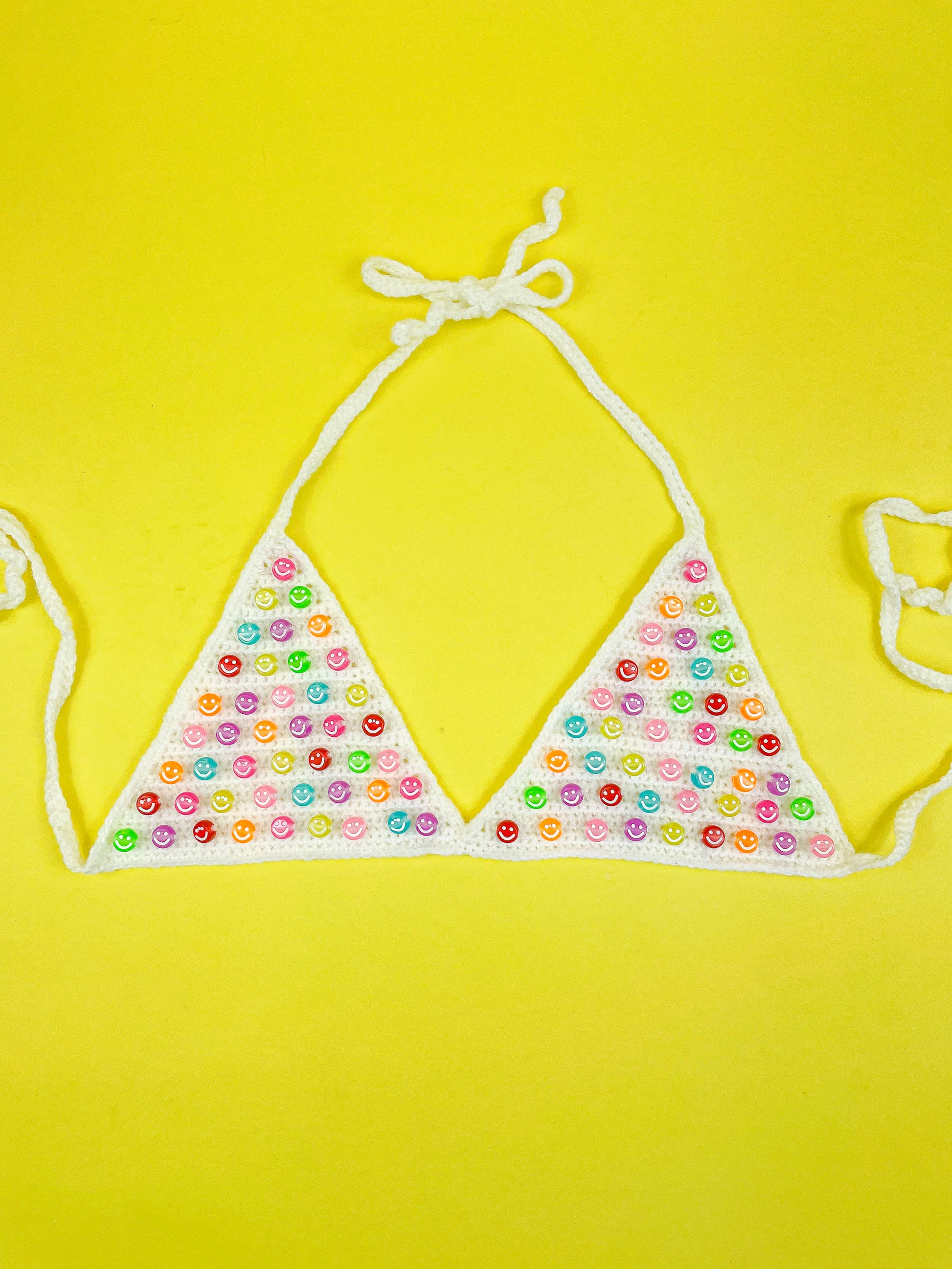 Flat lay of a white crochet bralette with multicoloured smiley face beads sewn into the cups lying on a yellow background.