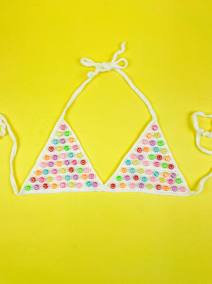 Flat lay of a white crochet bralette with multicoloured smiley face beads sewn into the cups lying on a yellow background.