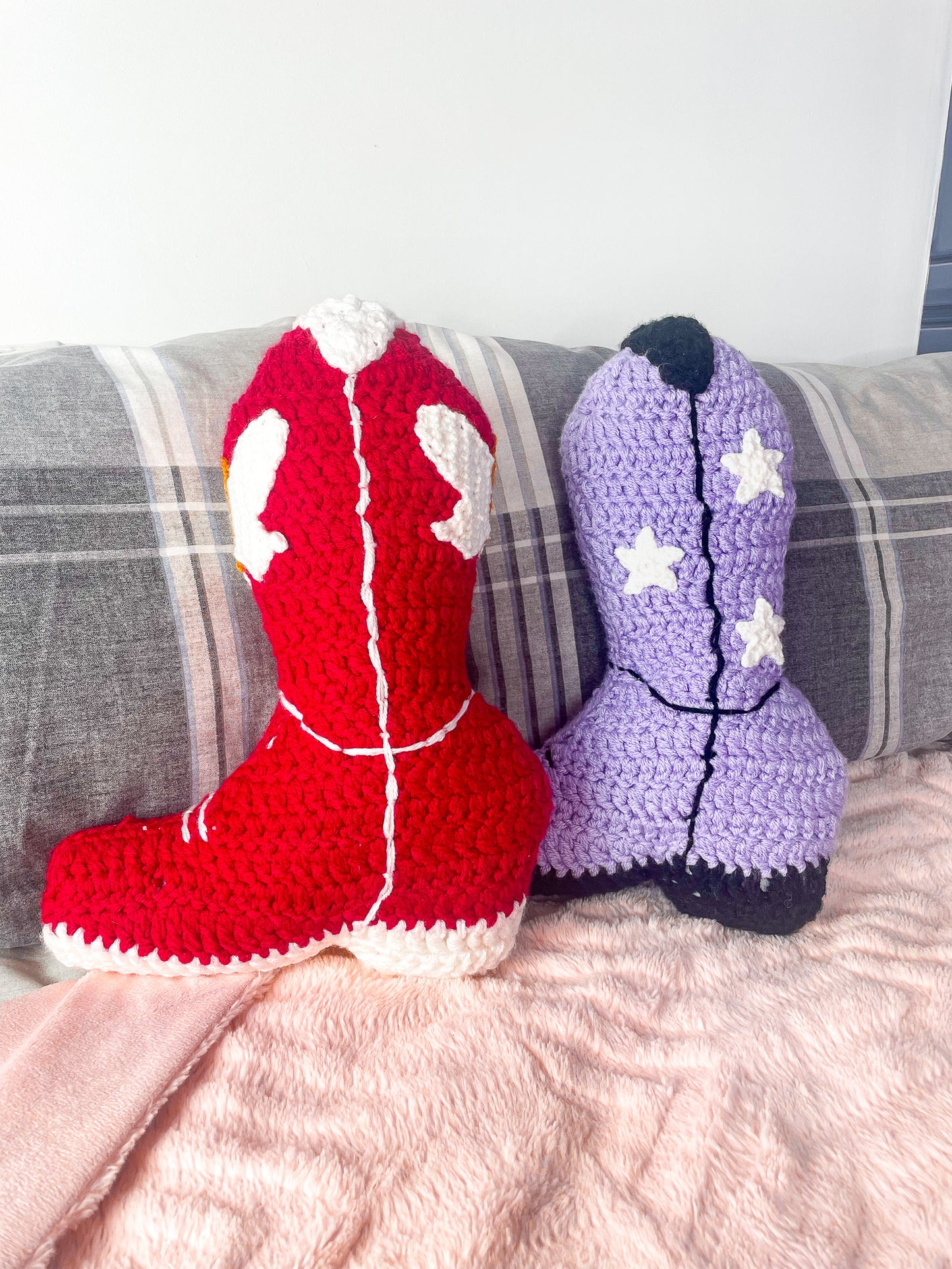 Two cowboy boot shaped cushions in red and lilac standing upright together leaning on two grey pillows on a bed with a pink blanket.