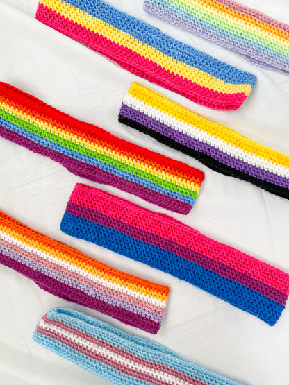 Seven crochet headbands in different pride flag colours (pastel rainbow, pansexual, non-binary, traditional, bisexual, lesbian, trans) lying on a white sheet.