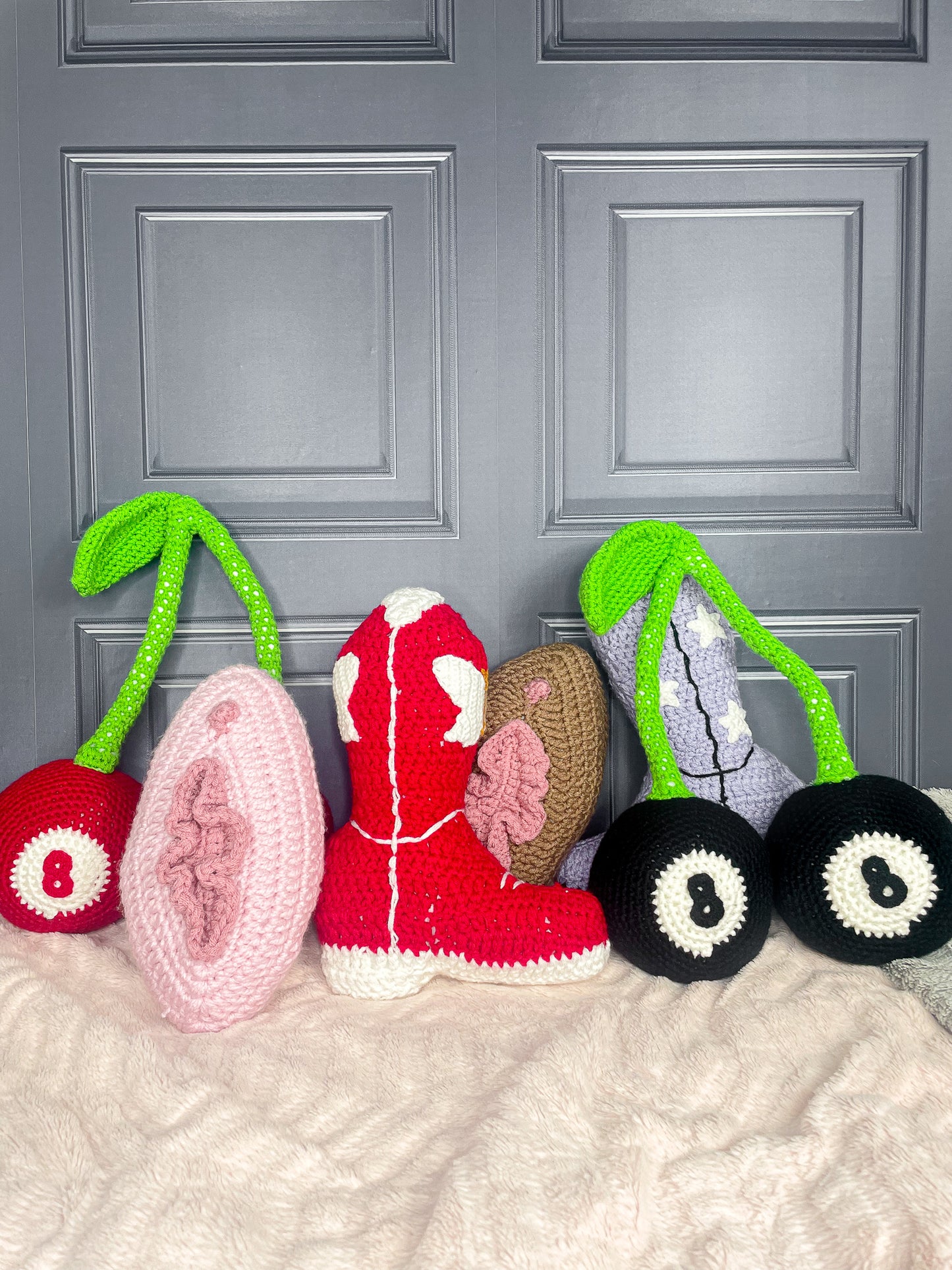 Six crochet cushions shaped like different things (two cherries, two cowboy boots, two vulvas), standing up right together against a grey wall.