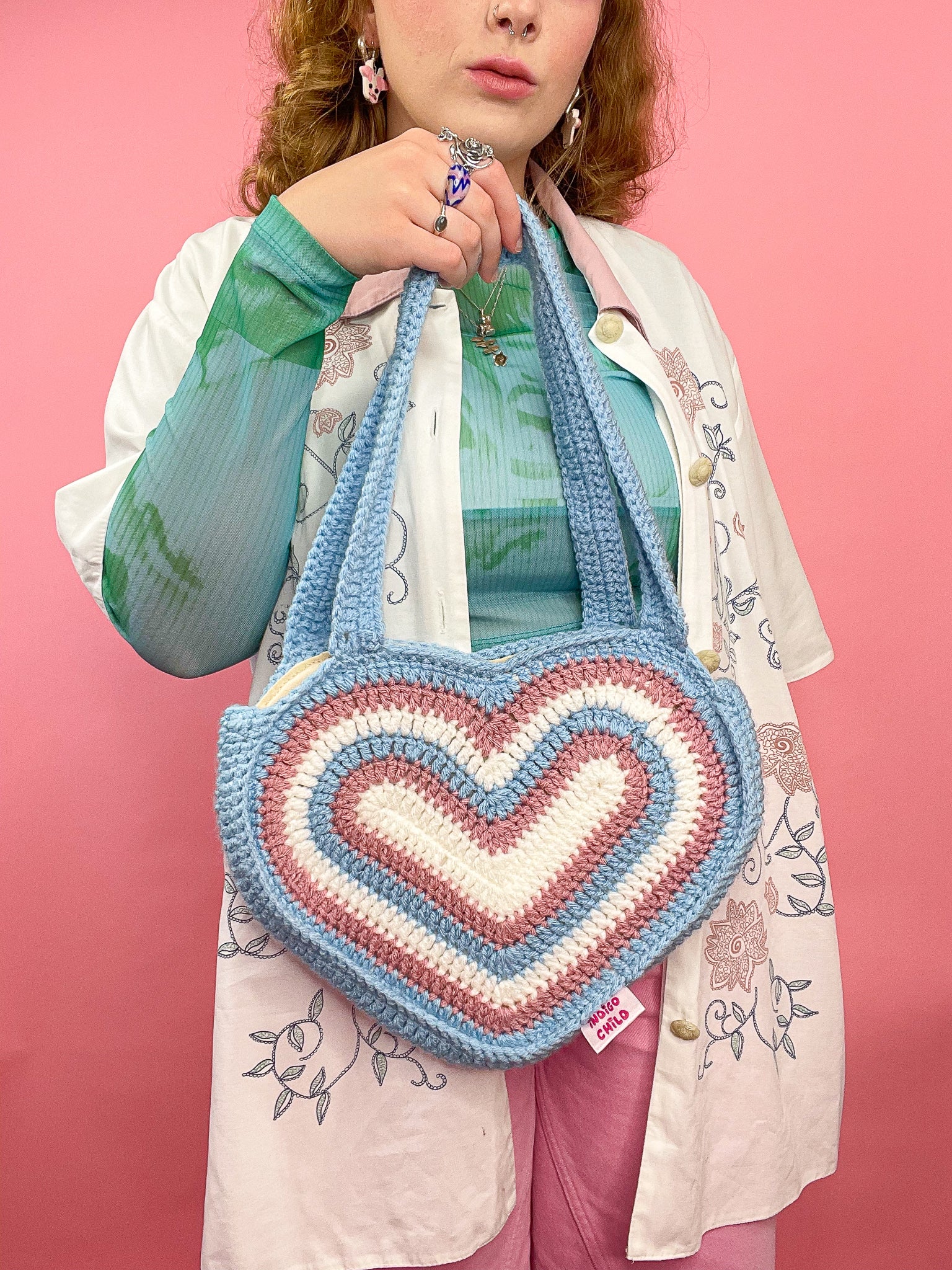 A woman holding a heart bag in the trans flag colours in front of her wearing a matching blue, pink and white outfit.