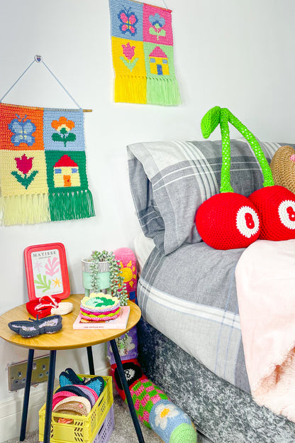 A bedroom set up with various crochet home furnishings (two wall hangings, a cherry shaped cushion, coasters, trinket dishes and a draught excluder) all placed around a room with other decor around.