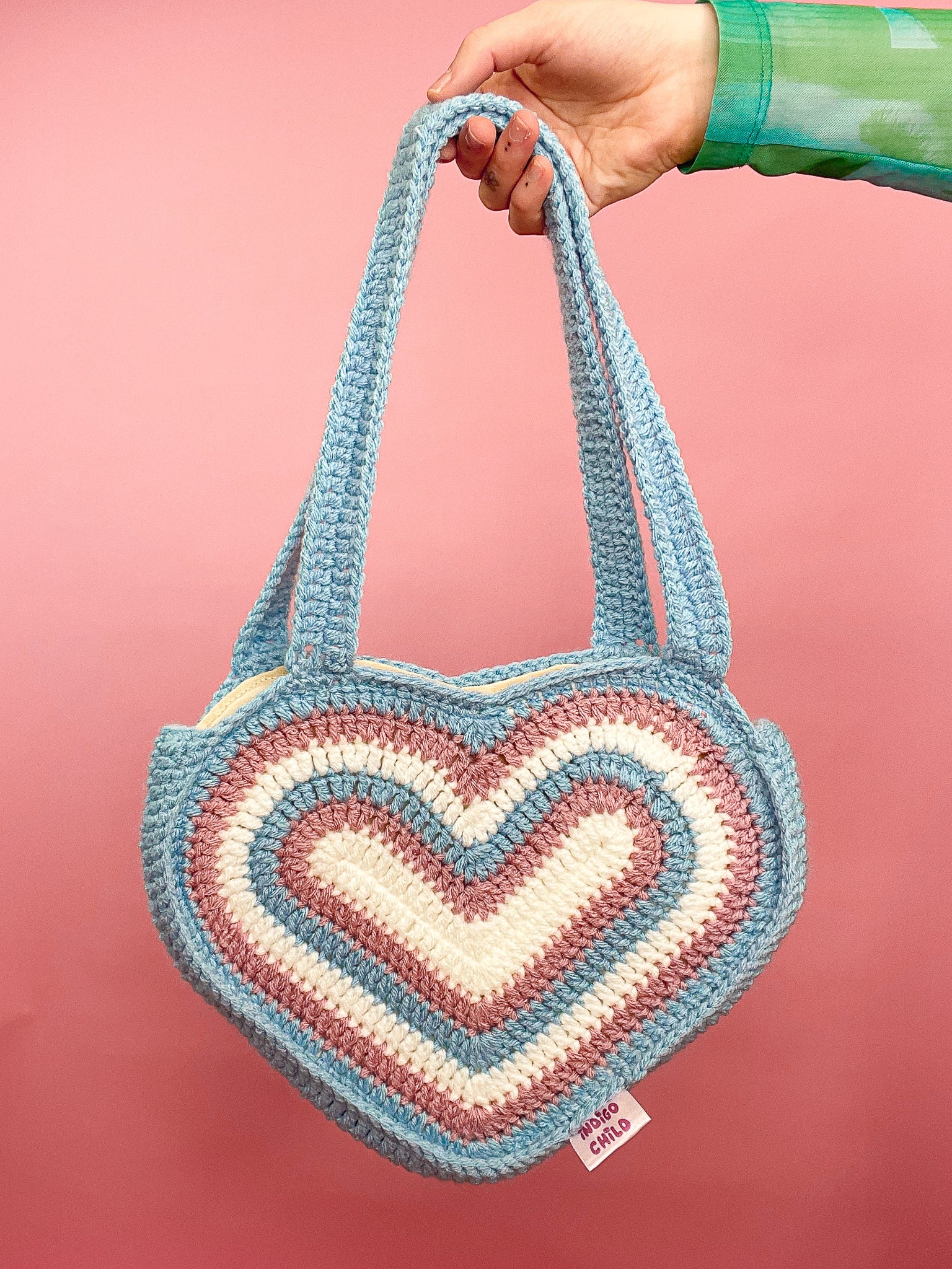 Close view of a hand holding a heart shaped bag in the trans flag colours (white, pink, blue) against a pink background.