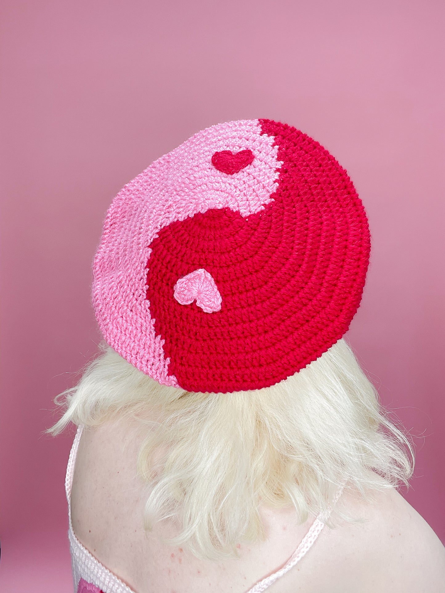 Back view of a woman wearing a pink and red crochet beret with a yin yang design and hearts for dots, in front of a pink background.