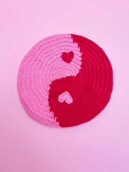 A crochet beret hat that looks like a yin and yang in pink and red, with appliqué hearts instead of dots.