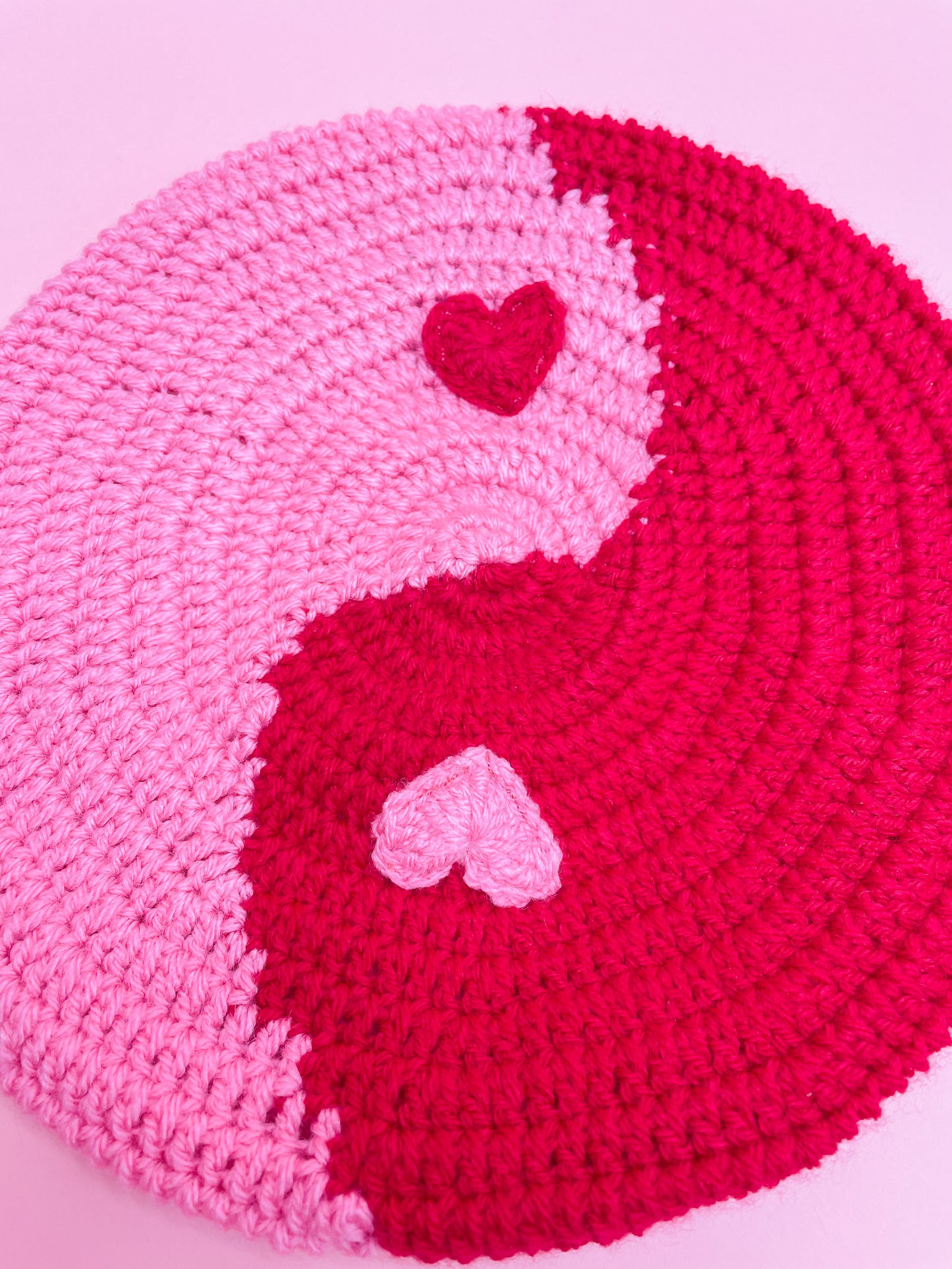 Close up of a crochet beret resembling a yin and yang symbol in pink and red, with hearts instead of dots.