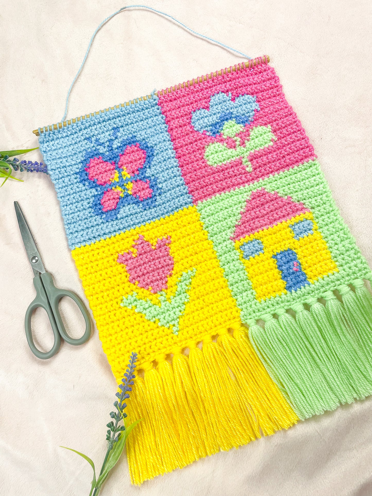 Flat lay of tapestry artwork in a pastel patchwork design (baby blue, pink, yellow, mint green), lying on a cream sheet with a pair of scissors and two lavender flowers around it.