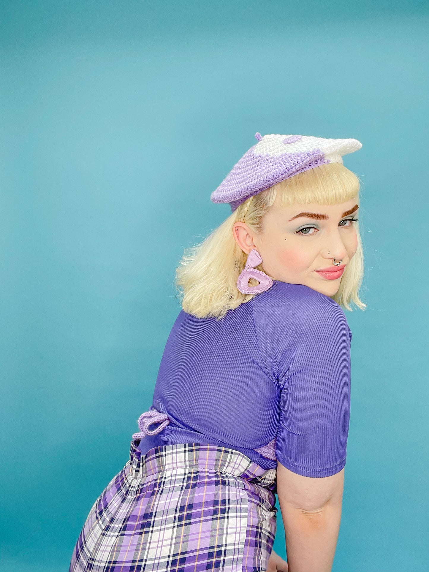 Side view of a woman posing wearing a crocheted beret in purple and white with a matching purple top, shorts and earrings.