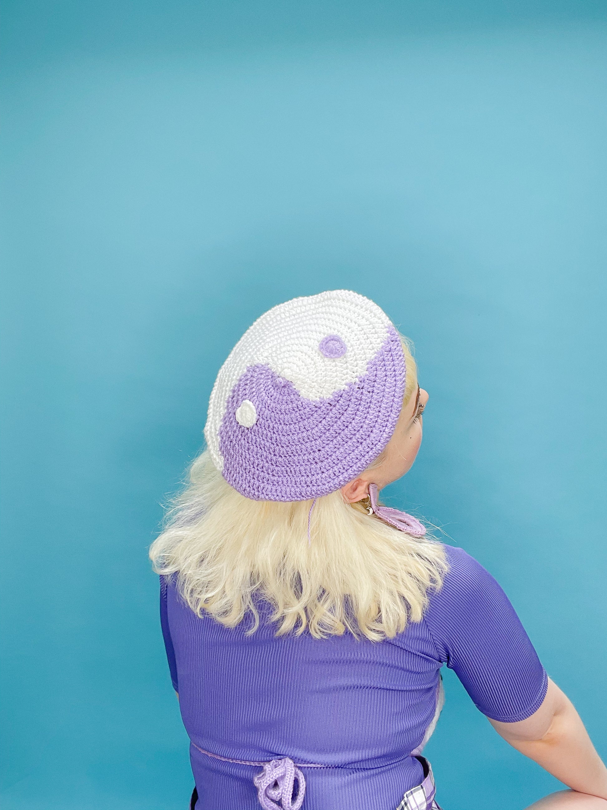 Back view of woman posing wearing a crochet yin and yang beret hat in purple and white with a matching purple top and earrings.