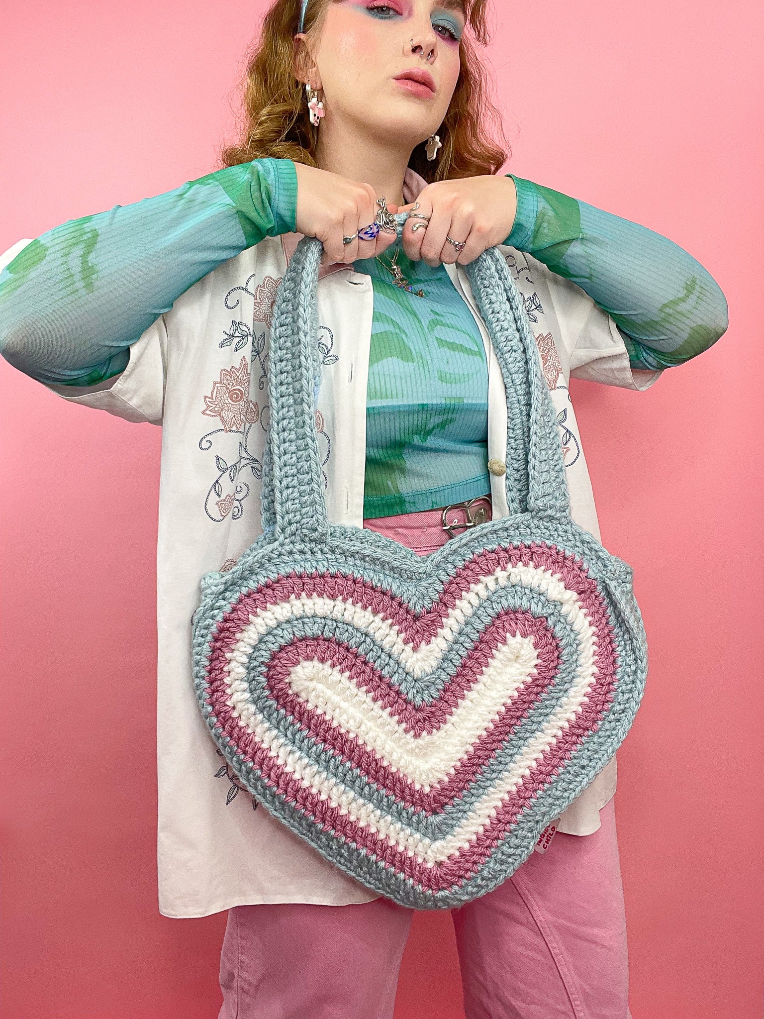 Close view of a woman posing holding a crochet heart bag in trans flag colours (white, pink and blue) paired with a matching outfit and makeup.