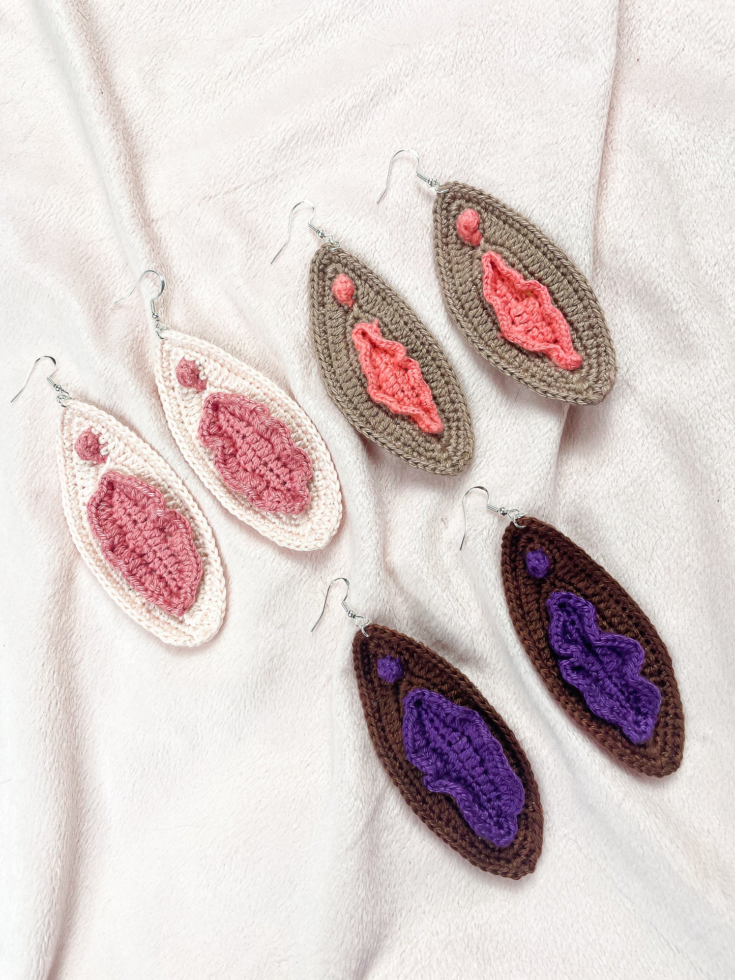 Three pairs of crochet vulva earrings in cream, light brown and dark brown lying flat on a cream background.