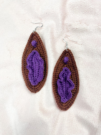 Flat lay of pair of dark brown vulva earrings with purple detailing with a 3D design for the clitoris and labia.