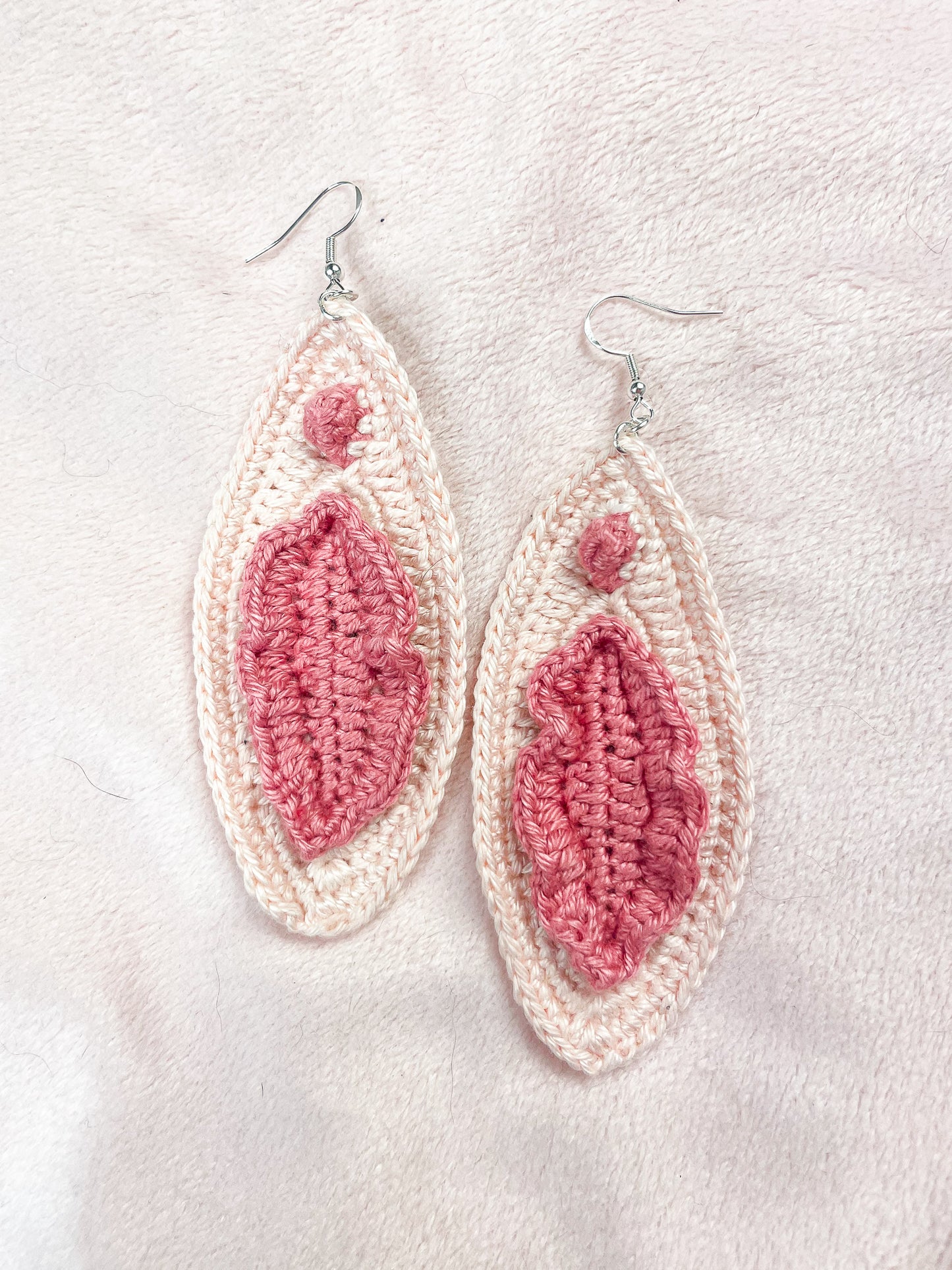 Flat lay of a pair of cream crochet earrings that look like vulvas lying on a pale pink background.