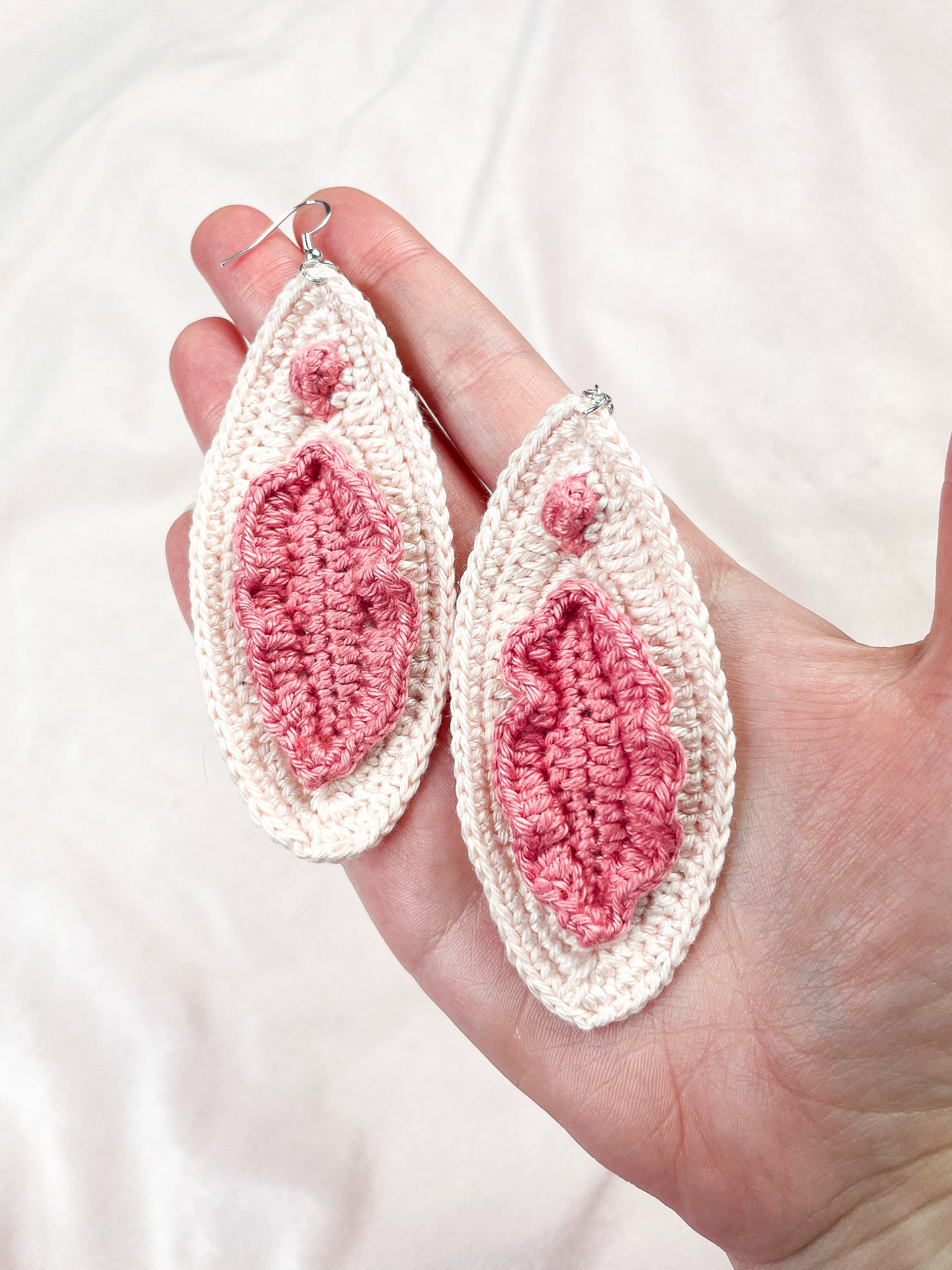 A hand holding a pair of cream crochet earrings with pink details showing the crochet stitches.