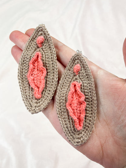 A hand holding a pair of crochet earrings resembling vulvas in brown with peach coloured details.