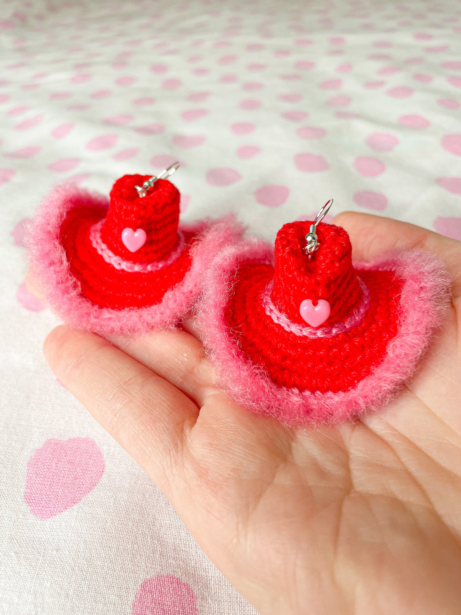 A hand holding a pair of red and pink crocheted earrings shaped like cowboy hats against a white and pink dotted background.