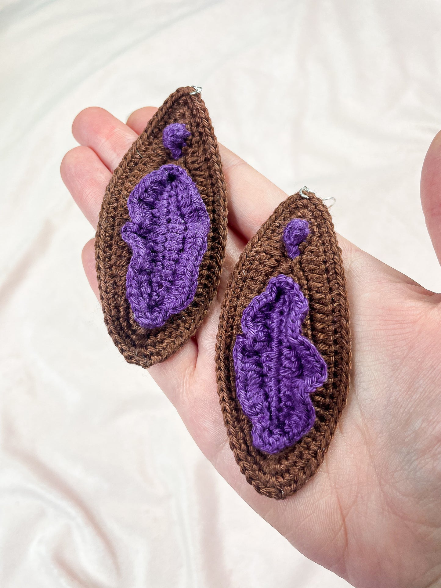 A hand holding a pair of vulva inspired earrings in dark brown with purple detailing.