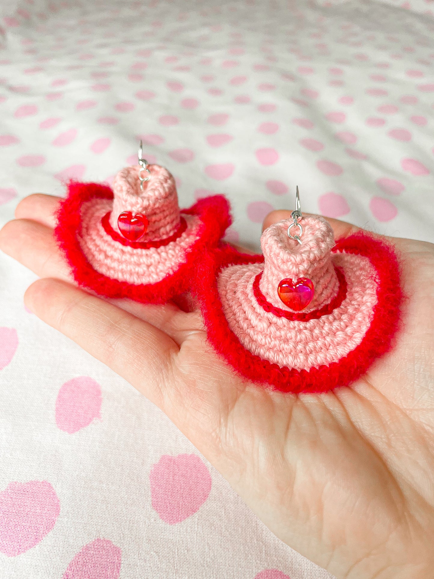 A hand holding a pair of crochet earrings shaped like cowboy hats in pink with red detailing in front of a white and pink dotted background.