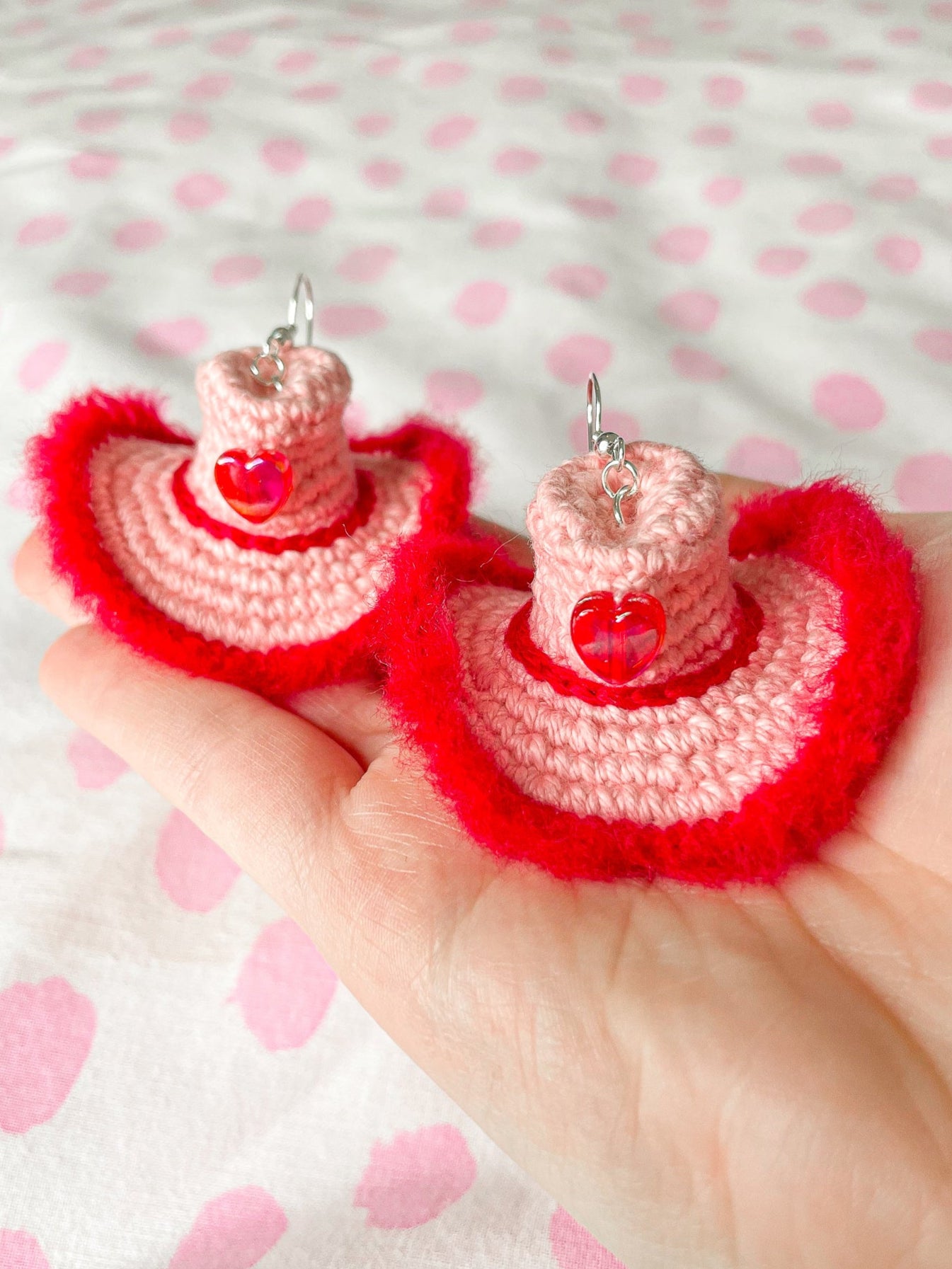 Close view of hand holding a pair of cowboy hat shaped earrings in pink with a fluffy red edge, red hatband and a heart bead at the front.