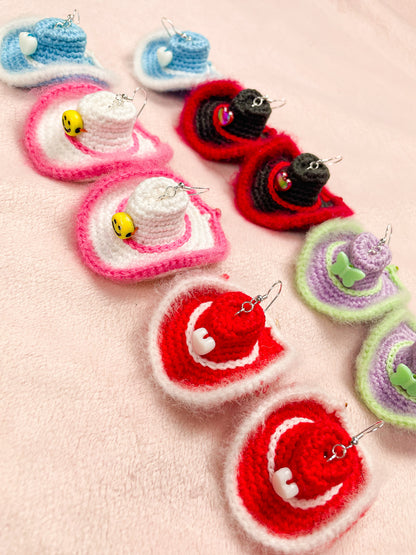 Five pairs of crochet earrings shaped like cowboy hats in multiple colours lying together in two rows on a pink background.