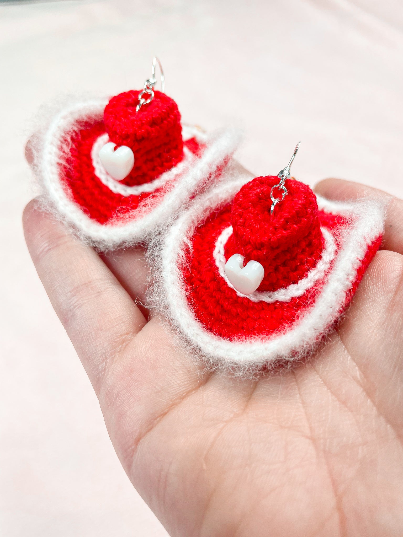 A hand holding a pair or red crochet earrings shaped like cowboy hats with white fluffy edges.