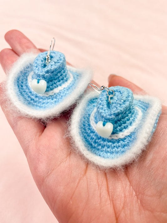 A hand holding a pair of cowboy hat shaped earrings in baby blue with white detailing.