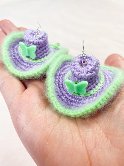 A hand holding a pair of crochet earrings shaped like cowboy hats in lilac with green details and a green butterfly bead on the front.